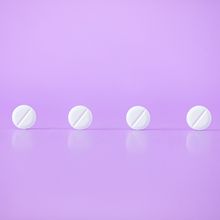 Four white, circular pills balance on their side in front of a pink background.