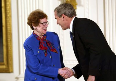 An older woman wearing a blue suit, shaking hands with an older man wearing a black suit.